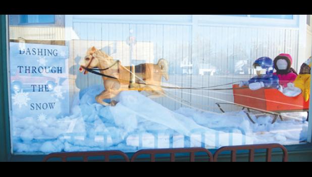 Mascot photo by Scott Thoma. Some of the local businesses have decorated their window fronts in winter and holiday scenes. With the sun shining bright on Saturday, reflections in the window cast a backdrop on this "Dashing Through the Snow" scene in the Public Library.